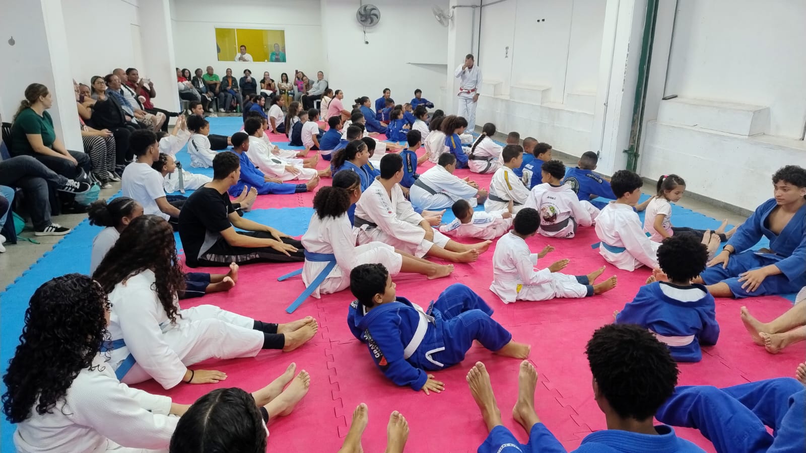Participantes no tatame, com familiares e acompanhantes ao redor.
